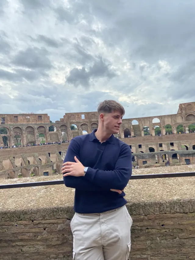 Leaning against a stone wall inside the Colosseum, looking away from the camera under a heavy sky.