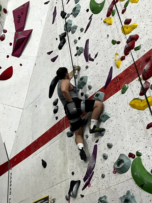 Climbing an indoor bouldering wall mid-route, roped in and reaching up for a hold, seen from behind and below.