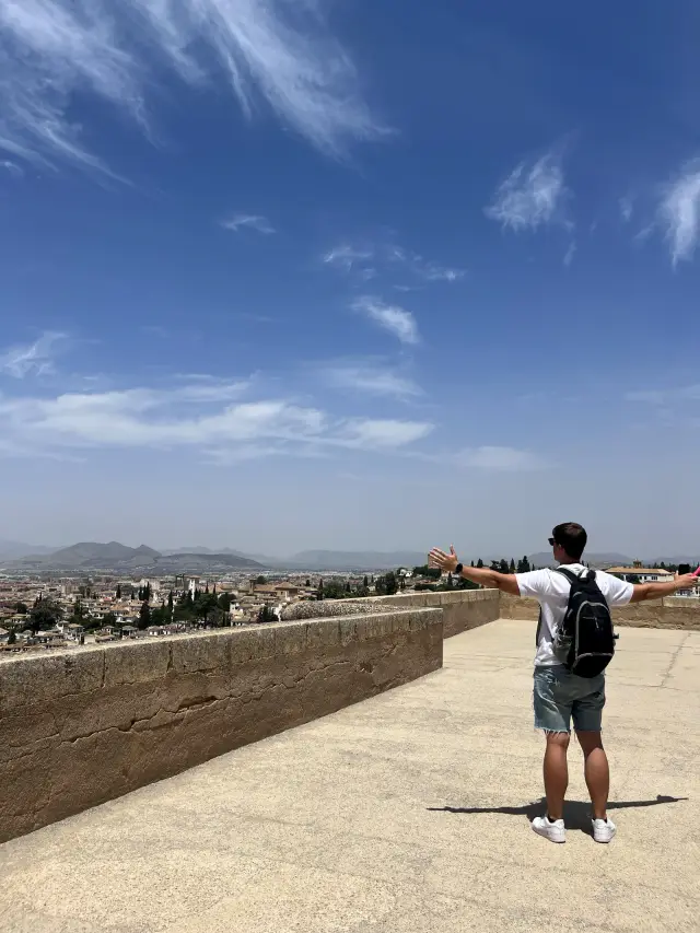 Standing on a fortress rampart above Granada, arms spread wide, overlooking the city and the mountains beyond under a bright blue sky.